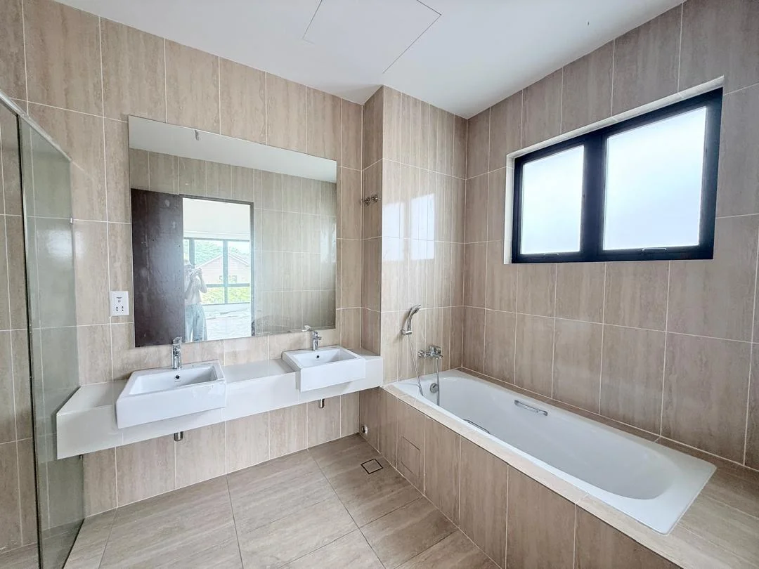 Bathroom features: double-sink vanity with white countertop, two chrome basin mixers, framed mirror above vanity, bathtub with white finish, shower area with glass partition, tile cladding on walls in beige, porcelain tile flooring in light beige, window with black frame, wall-mounted faucet, built-in shelving next to bathtub; style: modern.